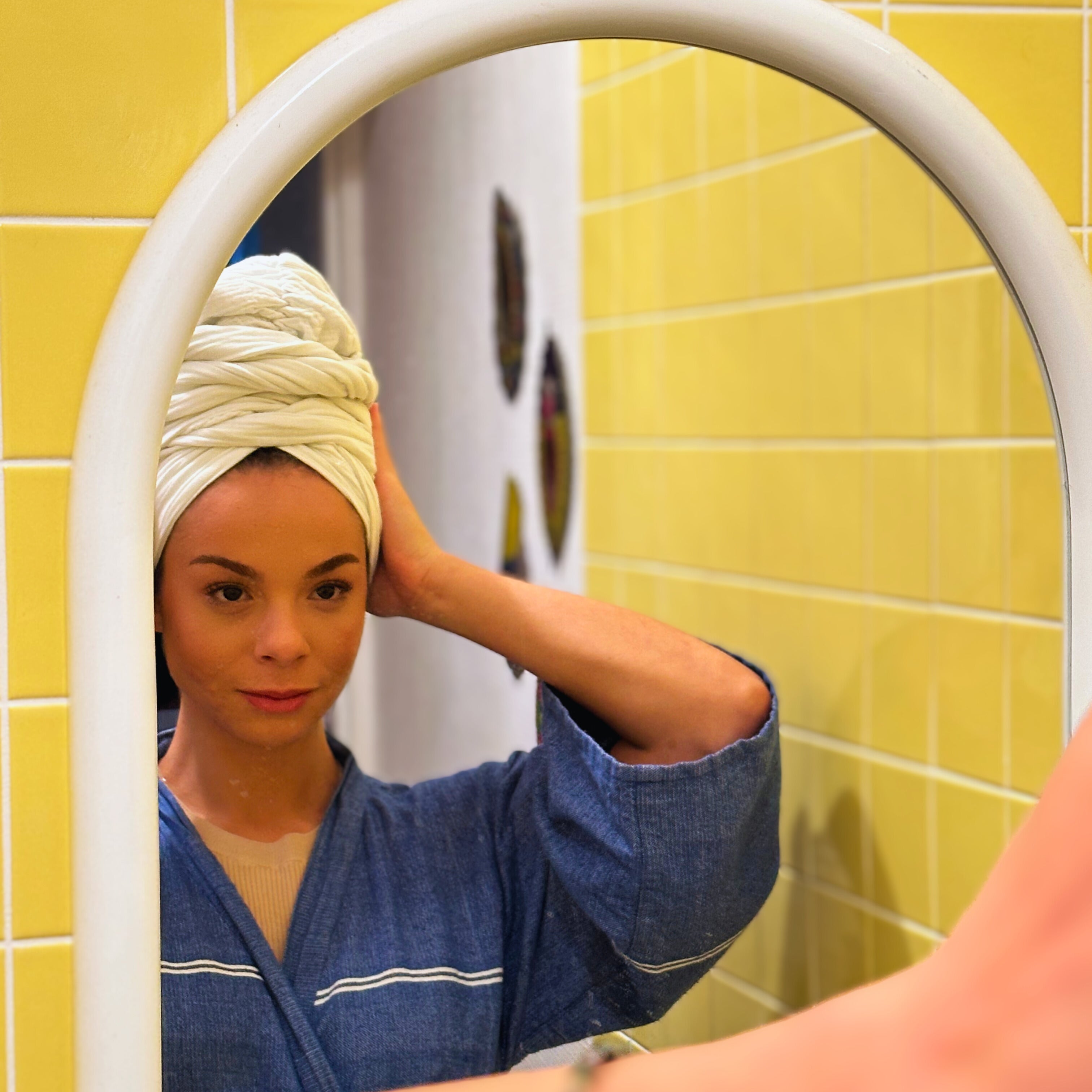 Woman wearing a white satin-lined turban, hands resting on the turban, looking at her reflection in the mirror