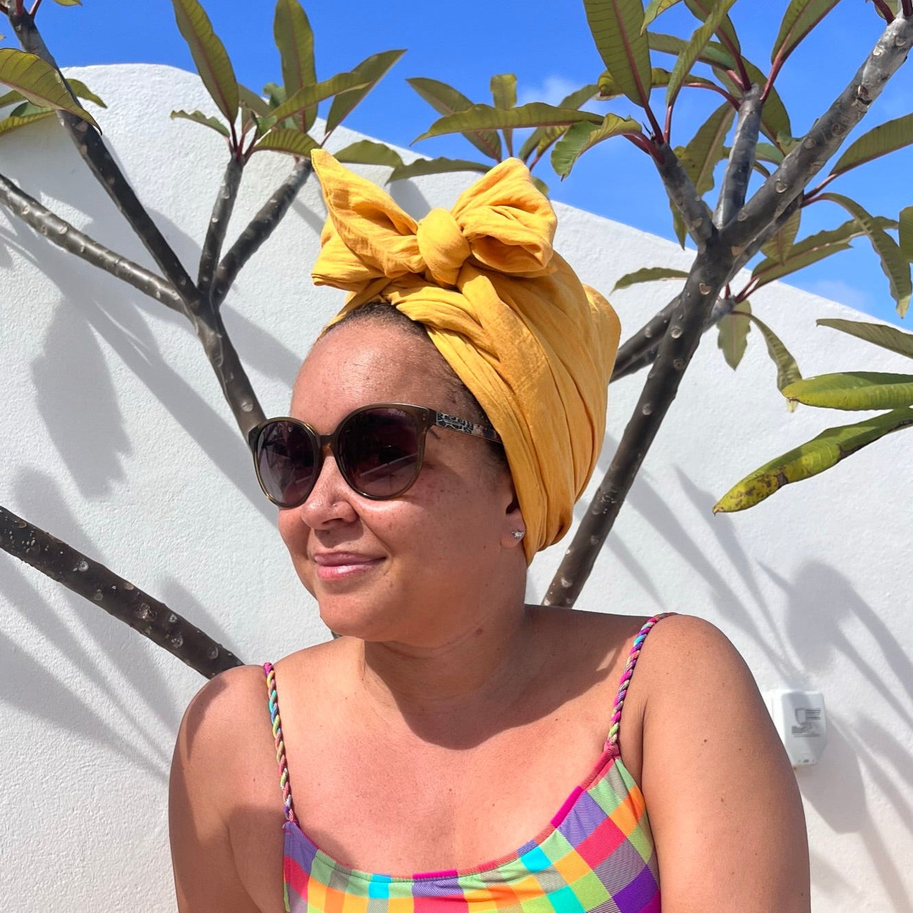 Woman wearing a yellow satin-lined turban, sunglasses and colourful top, sitting outside in the sunshine
