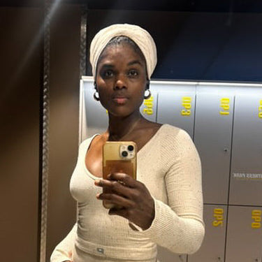 Woman wearing a white satin-lined turban, taking a mirror selfie on her phone in a gym locker room