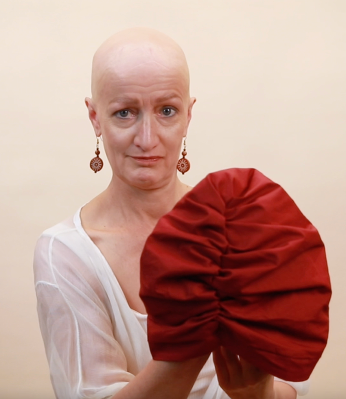 A woman with a bald head, holding up a red satin-lined turban in front of the camera, not yet wearing it