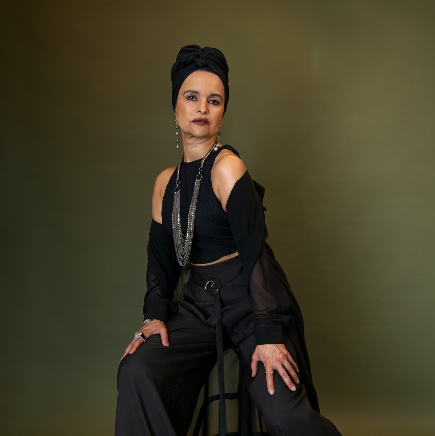 Woman wearing an all-black outfit, silver jewellery and a black turban, posed on a chair with her hands on her legs