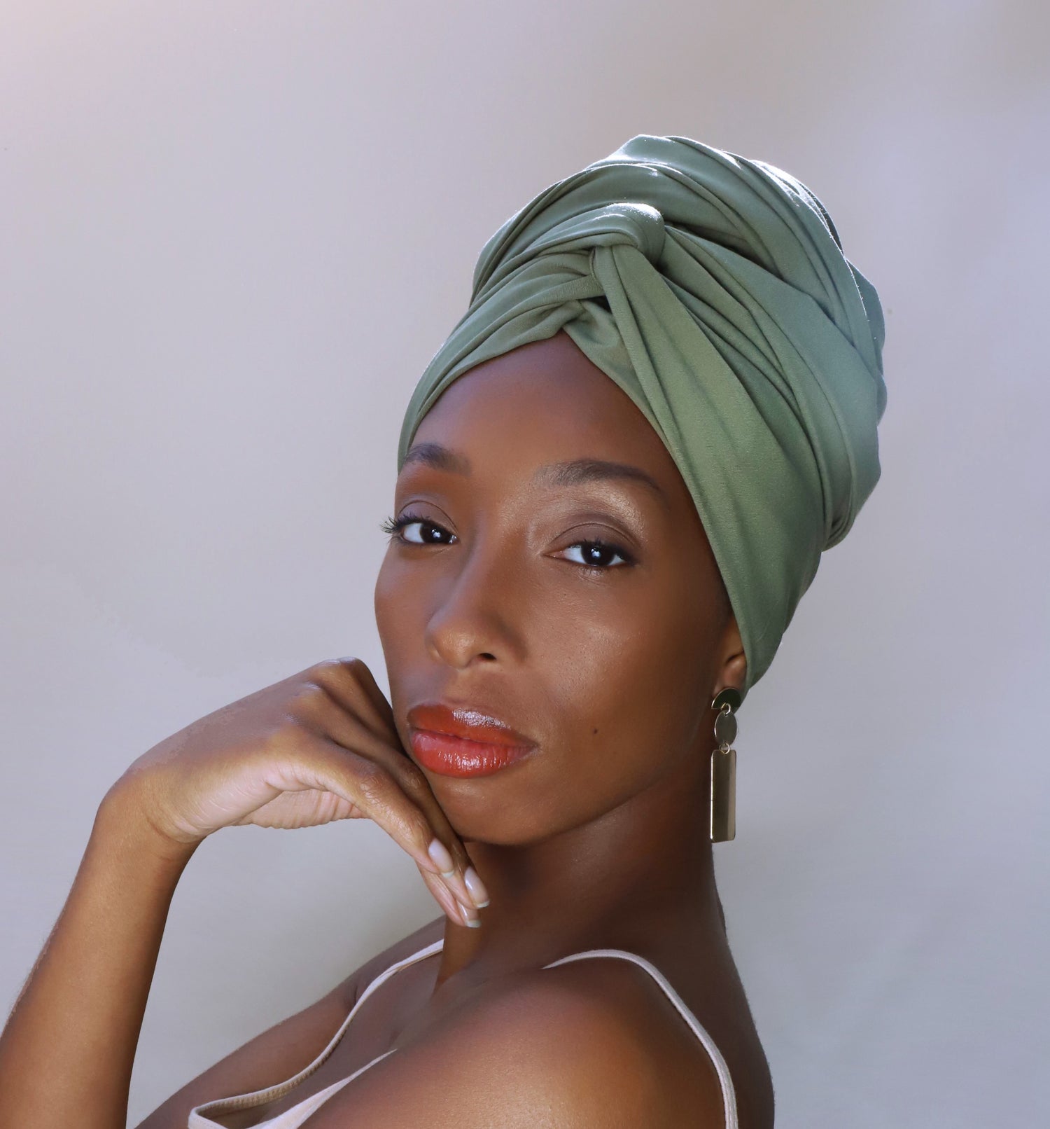 Woman wearing a sage green satin-lined turban, hand resting under her chin, looking over her shoulder at the camera
