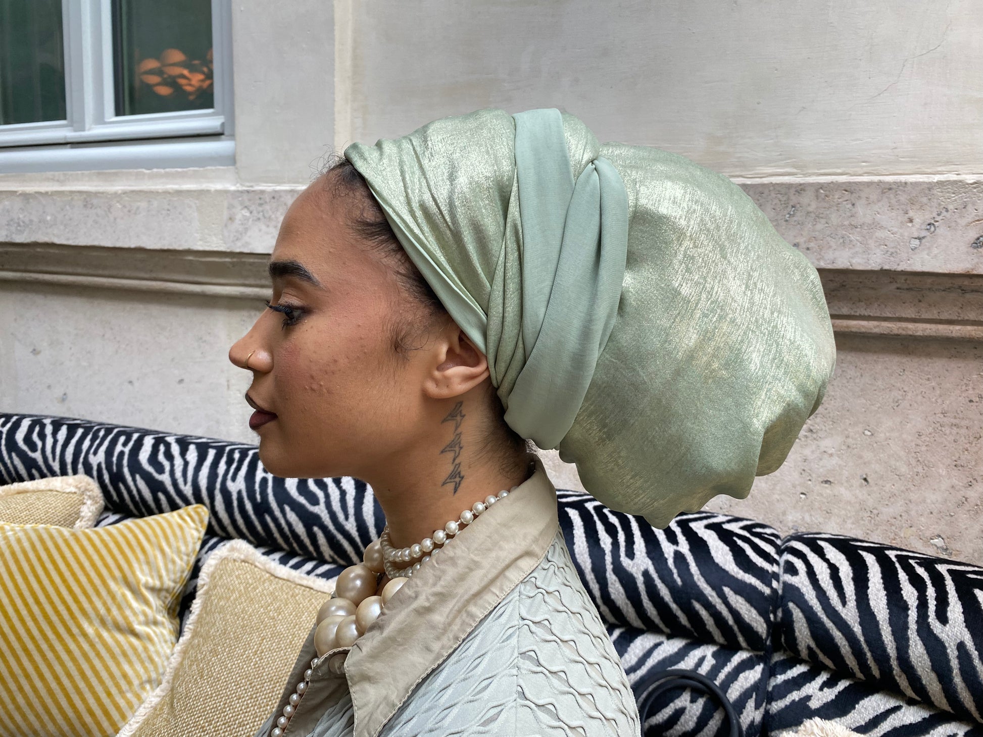 Side profile of a woman wearing a mint green metallic turban, seated outdoors near a window
