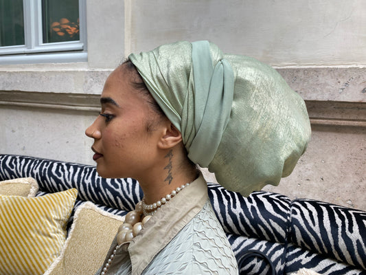 Side profile of a woman wearing a mint green metallic turban, seated outdoors near a window
